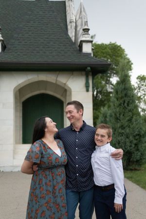 Sioux City, Iowa photographer. Family of 3 smiling and laughing together outside a charming white Gothic-style building with green trees.