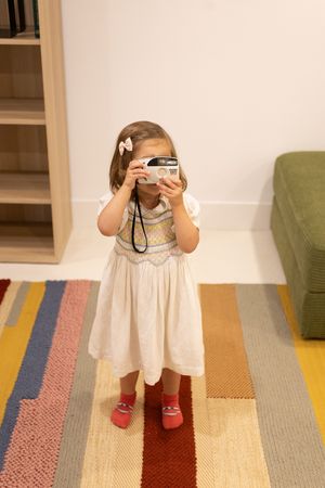 Sioux City, Iowa photographer. Cute toddler girl in white dress holding a film camera up to her face on a colorful striped rug.