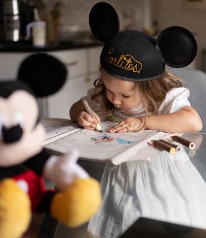 Sioux City, Iowa photographer. Young girl wearing Mickey Mouse ears hat colors at a table with a Mickey Mouse plush toy nearby.