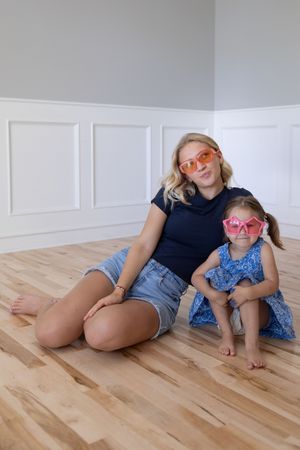 Sioux City, Iowa photographer. Blonde mother and young daughter in blue dress posing together on hardwood floor with pink sunglasses.