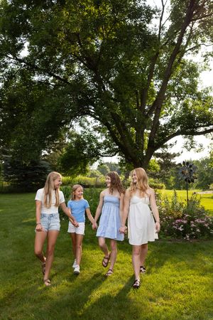 Sioux City, Iowa photographer. Four blonde girls in summer dresses holding hands and walking through a lush green garden on a sunny day.