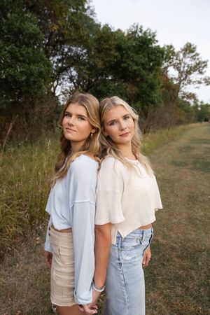 Sioux City, Iowa photographer. Two teenage girls stand back to back holding hands on a grassy trail lined with autumn trees.