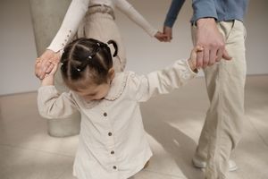 Sioux City, Iowa photographer. Toddler girl in white outfit holding hands with parents while playing in a bright minimalist indoor space.