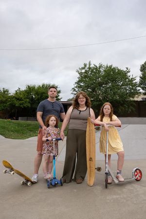 Family of four posing at a skate park with skateboards and scooters on a cloudy day.
