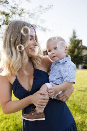 A smiling blonde woman in a navy dress holds a baby boy outdoors, surrounded by floating soap bubbles on a sunny day.