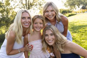Four smiling blonde women pose together outdoors on green grass, enjoying a sunny family portrait.