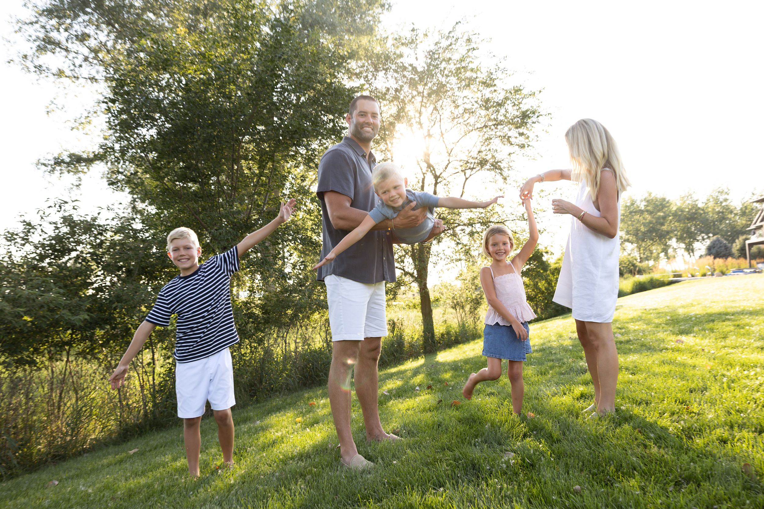Family of four playing outdoors on green grass in warm sunlight, with children spreading arms wide in joyful summer fun.