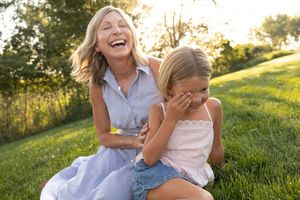 A laughing woman and a giggling young girl sit together on green grass in a sunny outdoor park setting.