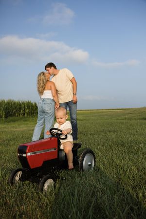 A baby sits on a red toy tractor in a green field while parents share a kiss behind them.