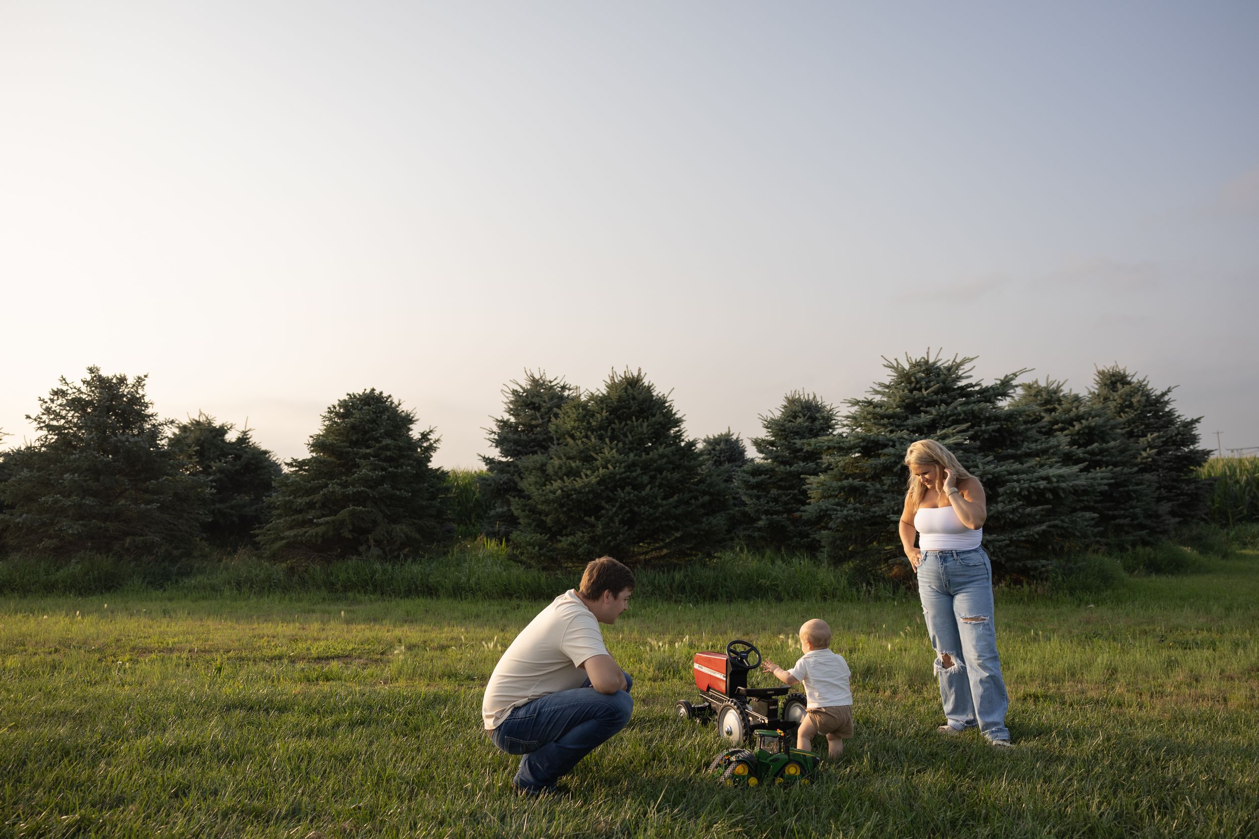 Family with toddler playing in a green meadow with evergreen trees in the background at dusk.