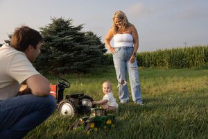 Family outdoors with baby sitting on toy John Deere tractor in grassy field as parents watch and photograph.