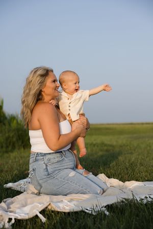 Smiling blonde mother holding happy baby on picnic blanket in green field under blue sky.