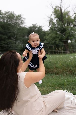 A mother sits on a blanket outdoors, lifting her smiling baby girl in a navy sailor dress up into the air.