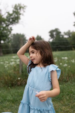 Young girl in a blue ruffle dress poses outdoors in a green field, hand raised to her head.