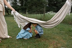 Two children hiding under a fringed hammock in a lush green backyard while an adult holds it up.