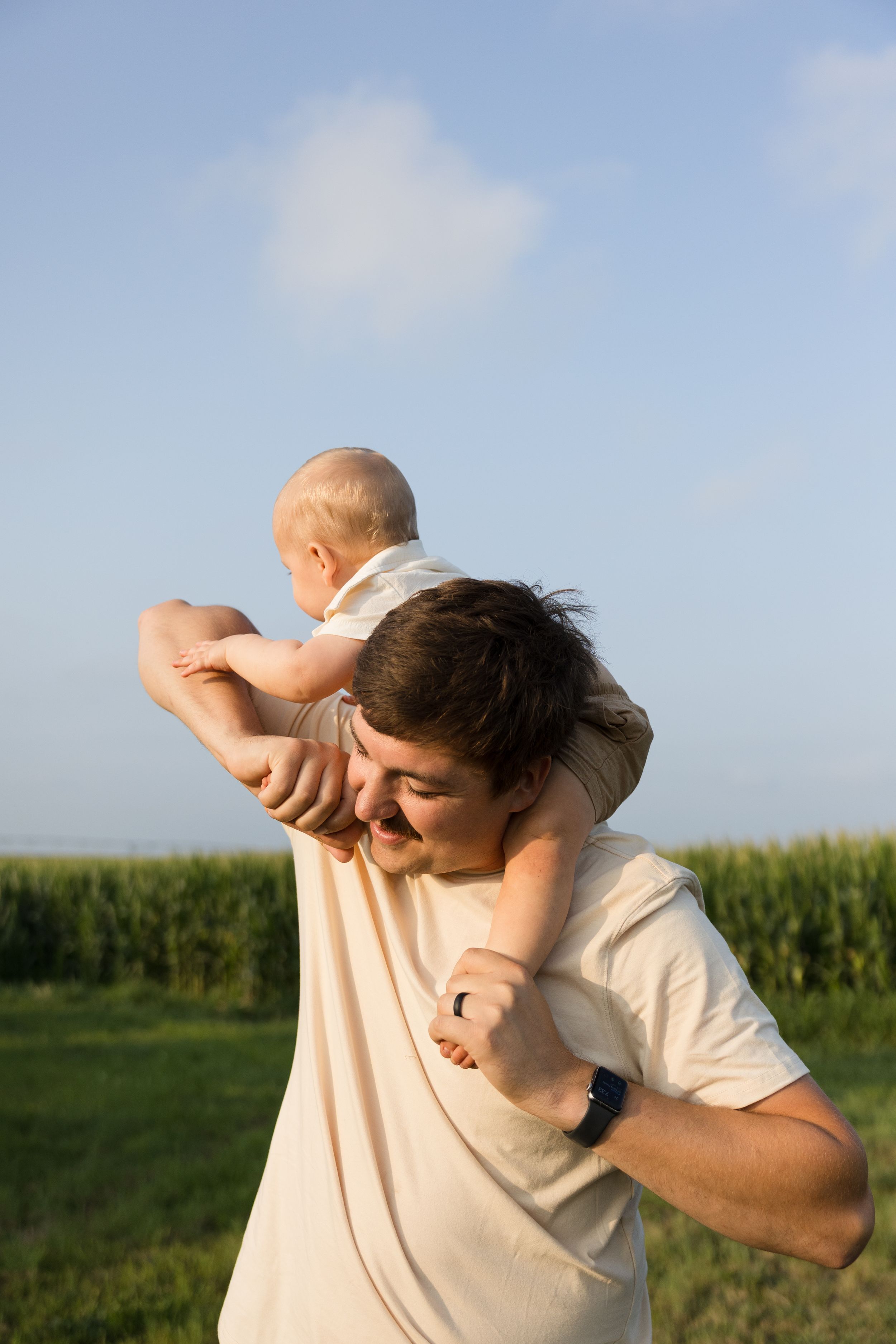 Father carrying baby on shoulders in sunny field with green corn crops and blue sky in background.