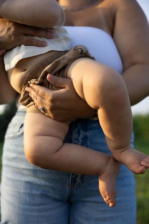 Mother holding chubby baby outdoors in warm sunlight, baby wearing brown outfit with tiny bare feet visible.