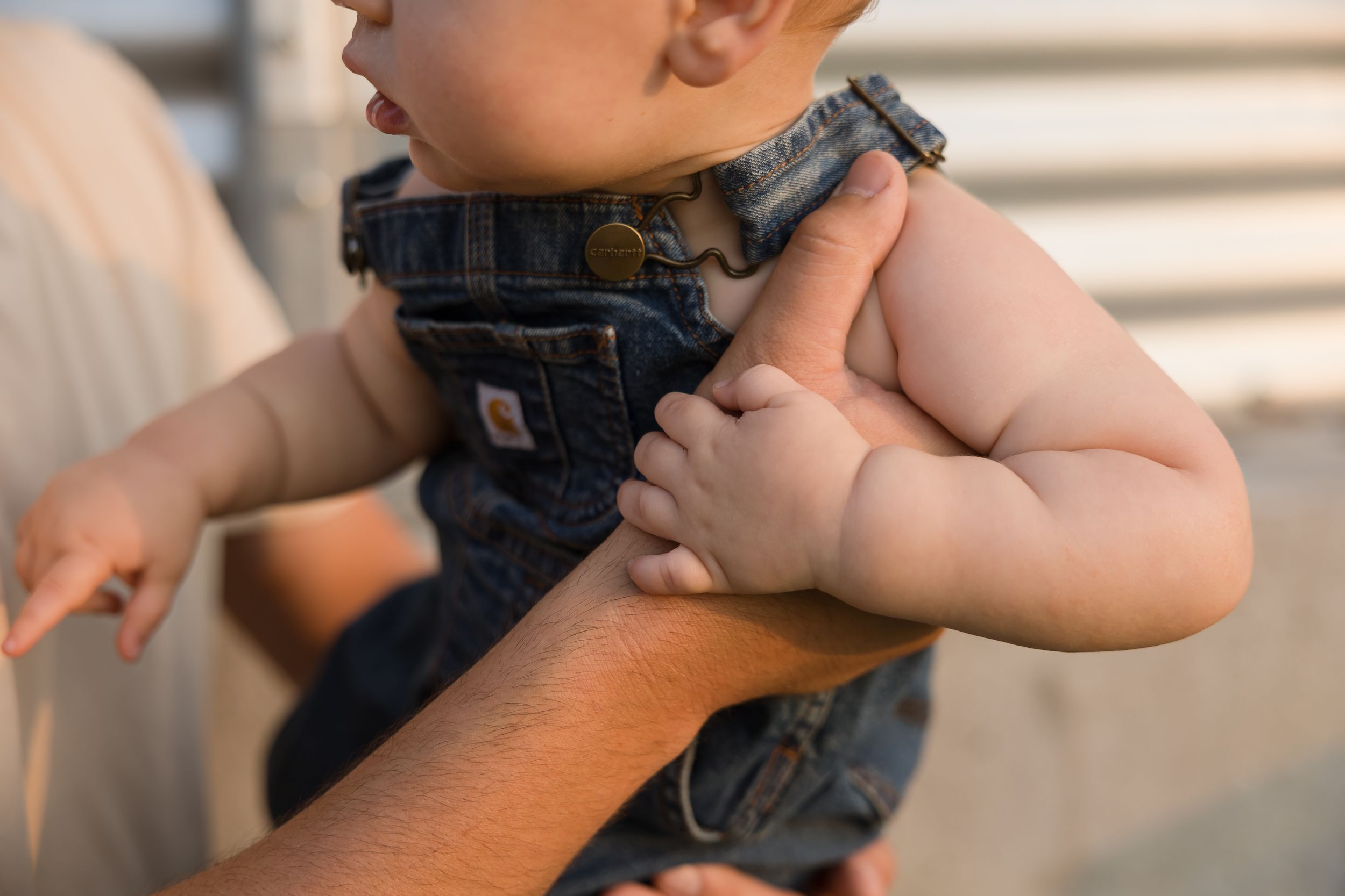 A baby in denim overalls being held by an adult, with chubby hands gripping the caregiver's arm.