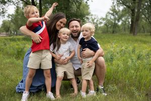 Happy family of five smiling outdoors in a lush green park, parents holding young children on a sunny day.