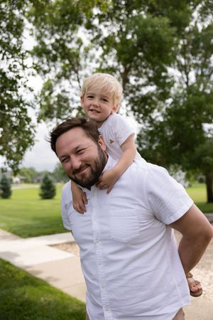 A bearded man in a white shirt carries a smiling blonde toddler on his shoulders in a lush green park.