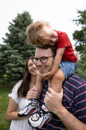 A happy family outdoors, with a young child sitting on dad's shoulders while mom smiles beside them near evergreen trees.