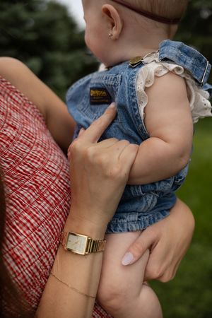 Mother holding baby girl in denim romper, wearing gold watch and red plaid dress, outdoors.