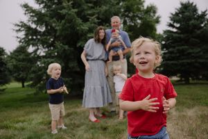 A joyful family poses outdoors on green grass with tall trees, as a young boy in a red shirt laughs in the foreground.
