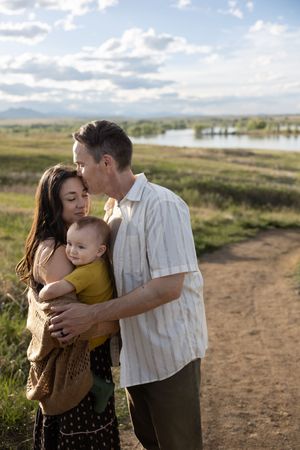 A family of three shares a tender moment outdoors, with parents embracing their baby near a scenic lake.
