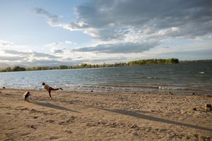 Person playing fetch with a dog on a sandy beach beside a calm lake under a dramatic cloudy sky.