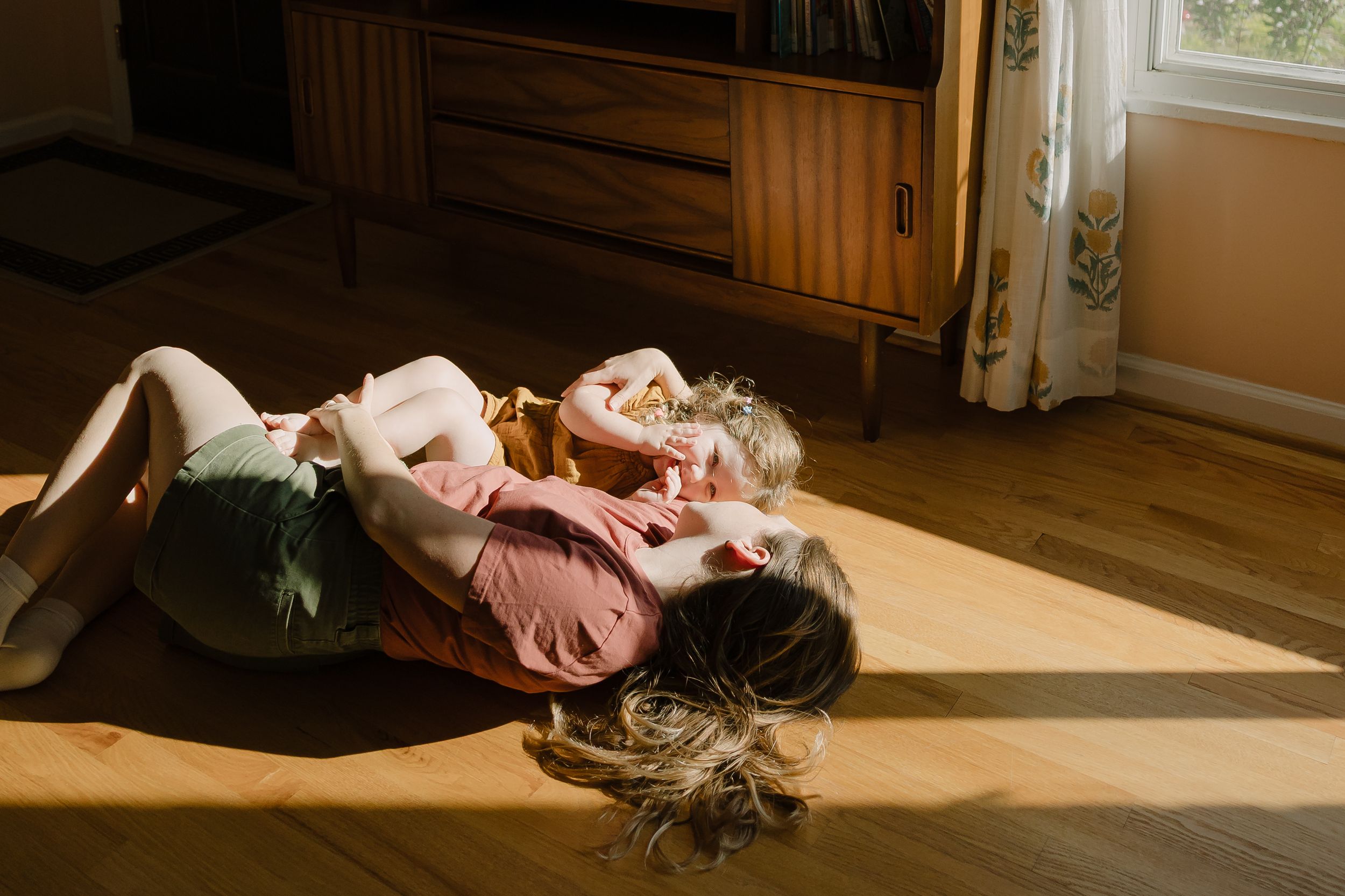 Sioux City, Iowa photographer. Mother and child cuddling on a sunlit wooden floor in a cozy living room.