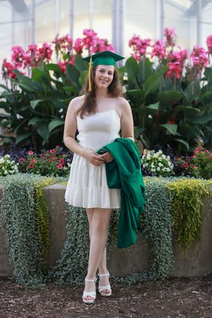 A person in a white dress poses in front of pink orchids and greenery for graduation photos.