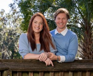 Couple poses together on rustic wooden railing outdoors with palm trees in background.