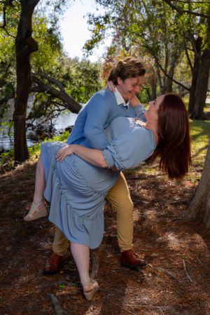 Playful couple dances outdoors on pine needle covered ground near waterfront.