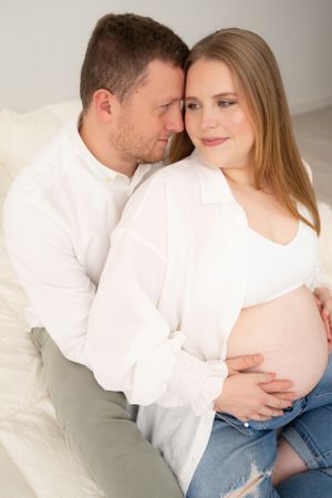 Intimate maternity photoshoot with couple in white shirts and jeans against light background.