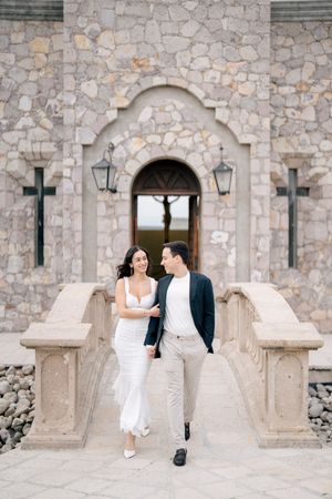 Romantic couple in wedding attire sits together on stone benches against a rustic stone church wall with arched doorways.