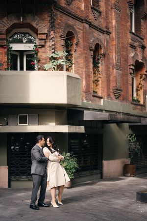 Two people share a tender moment outside a historic brick building with modern architectural details.