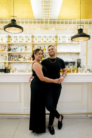 A couple in all black attire embraces while standing at an elegant bar with gold accents and pendant lighting.