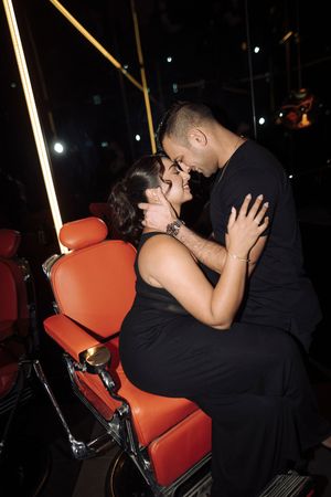 A couple dressed in black share an intimate moment while seated on red barber chairs at nighttime.