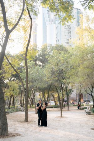 Two people in long black dresses stand together on a tree-lined path in an urban park with tall buildings visible beyond.