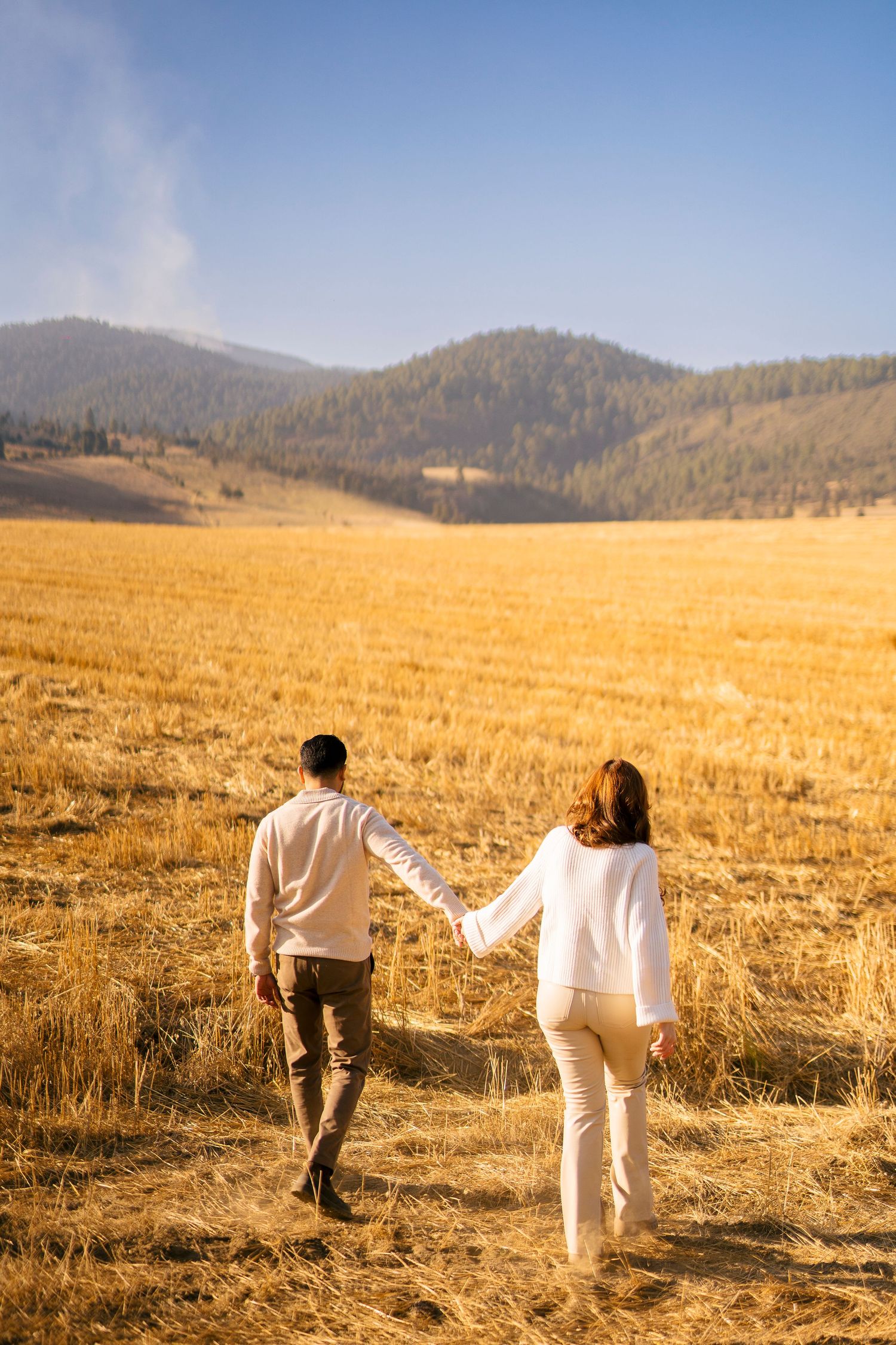 Couple walking hand in hand through golden wheat field with mountains in background.