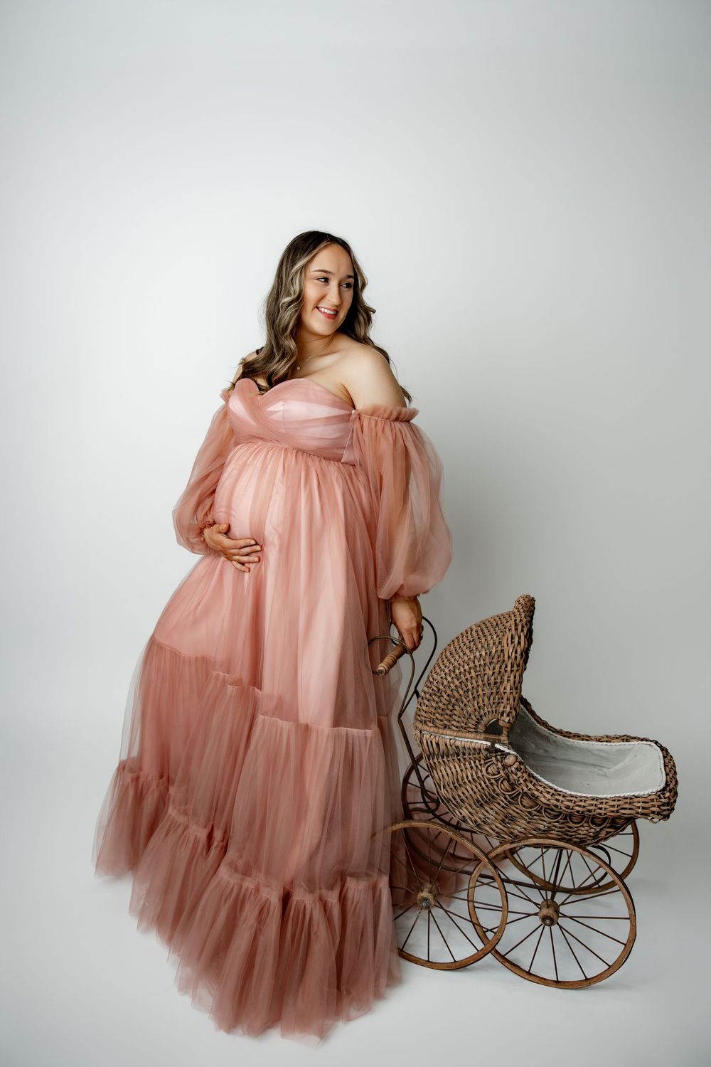 A person in a flowing pink tulle maternity gown poses elegantly next to a vintage wicker baby carriage against a white backdrop.