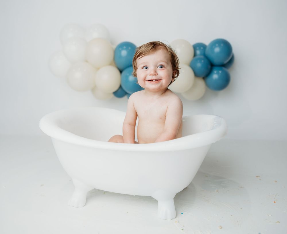 A happy baby sits in a white clawfoot bathtub with blue and white balloons decorating the background.