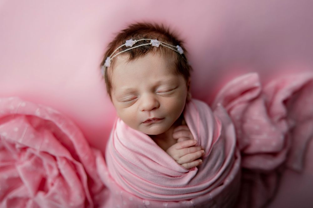 Sleeping newborn baby wrapped in soft pink fabric against pink background during professional photoshoot.