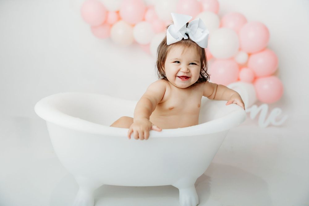 Baby sitting in white bathtub with pink balloon arch backdrop for milestone photo session.