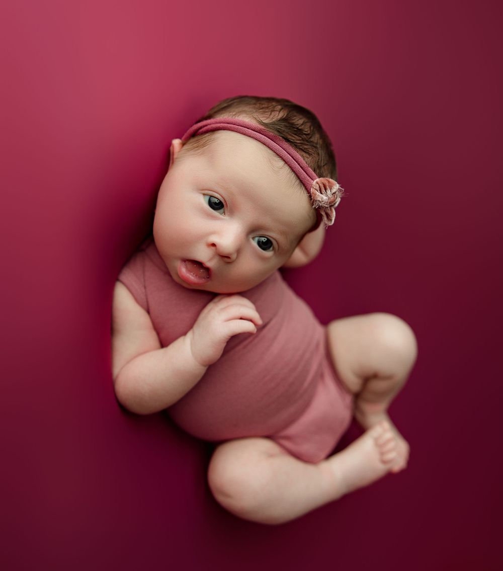 Newborn in pink outfit lies on burgundy backdrop during professional portrait session.