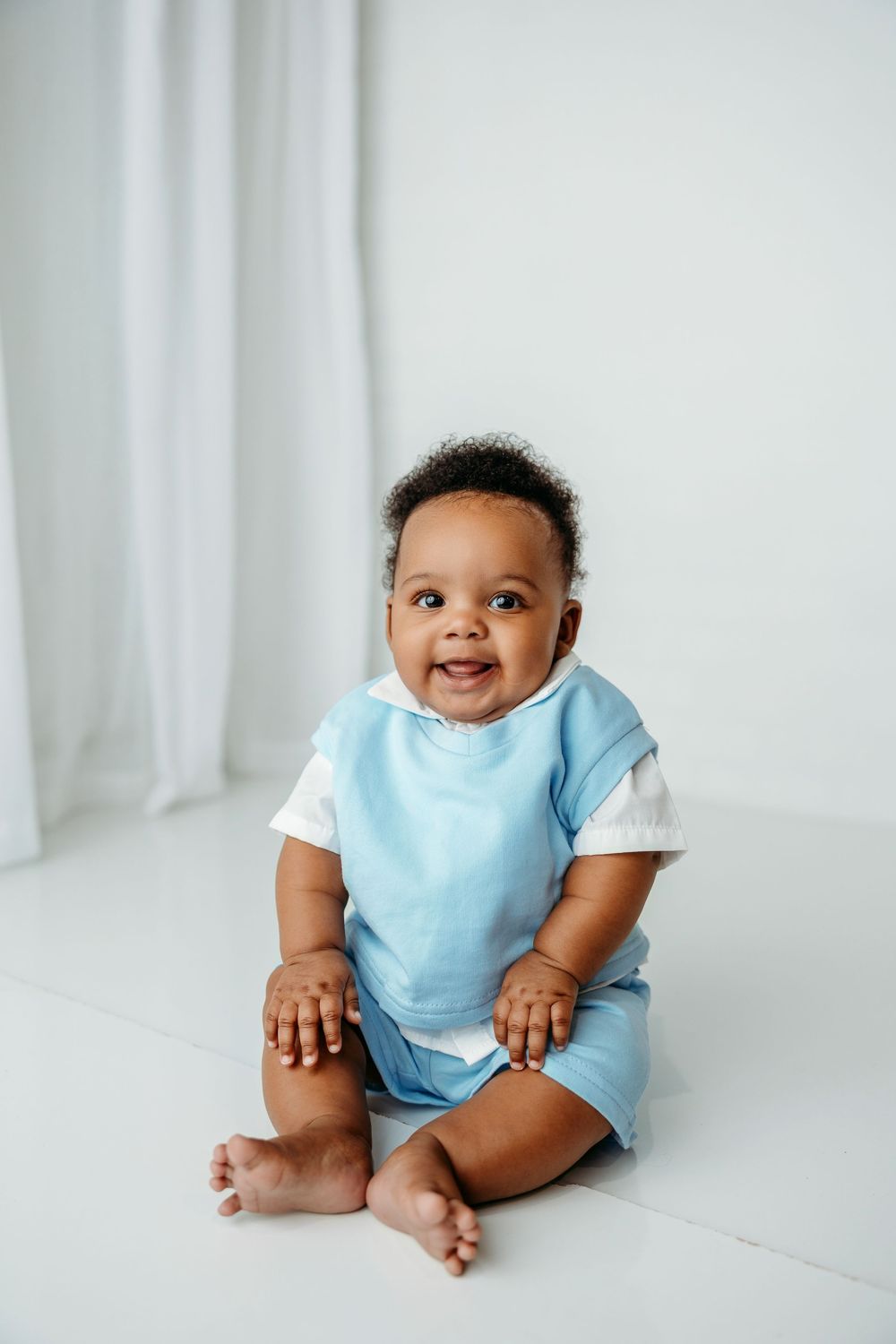 A happy baby in a light blue onesie sits on a white surface against a white backdrop, creating a minimalist portrait.