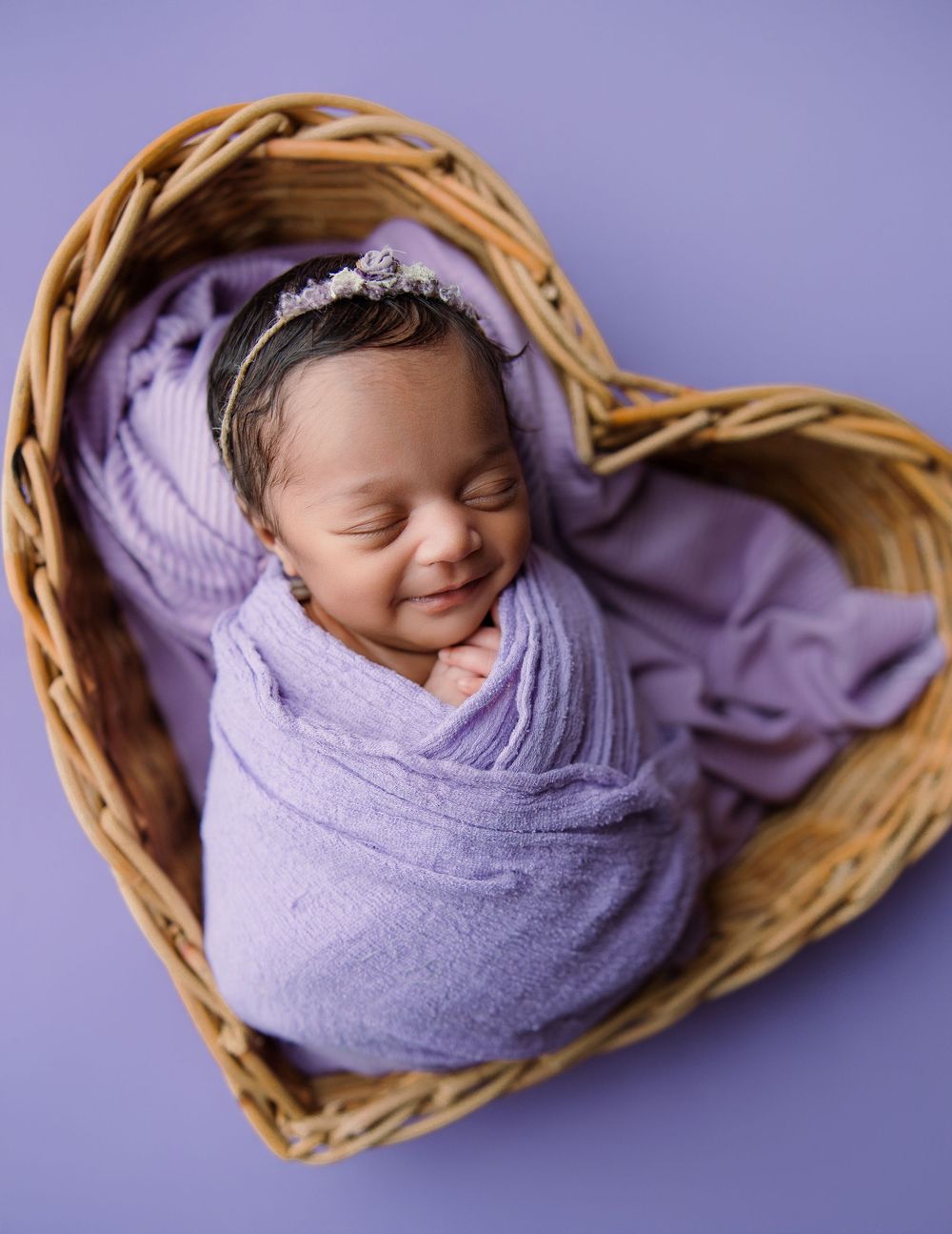Sleeping newborn wrapped in purple swaddle rests peacefully in heart-shaped wicker basket against purple background.