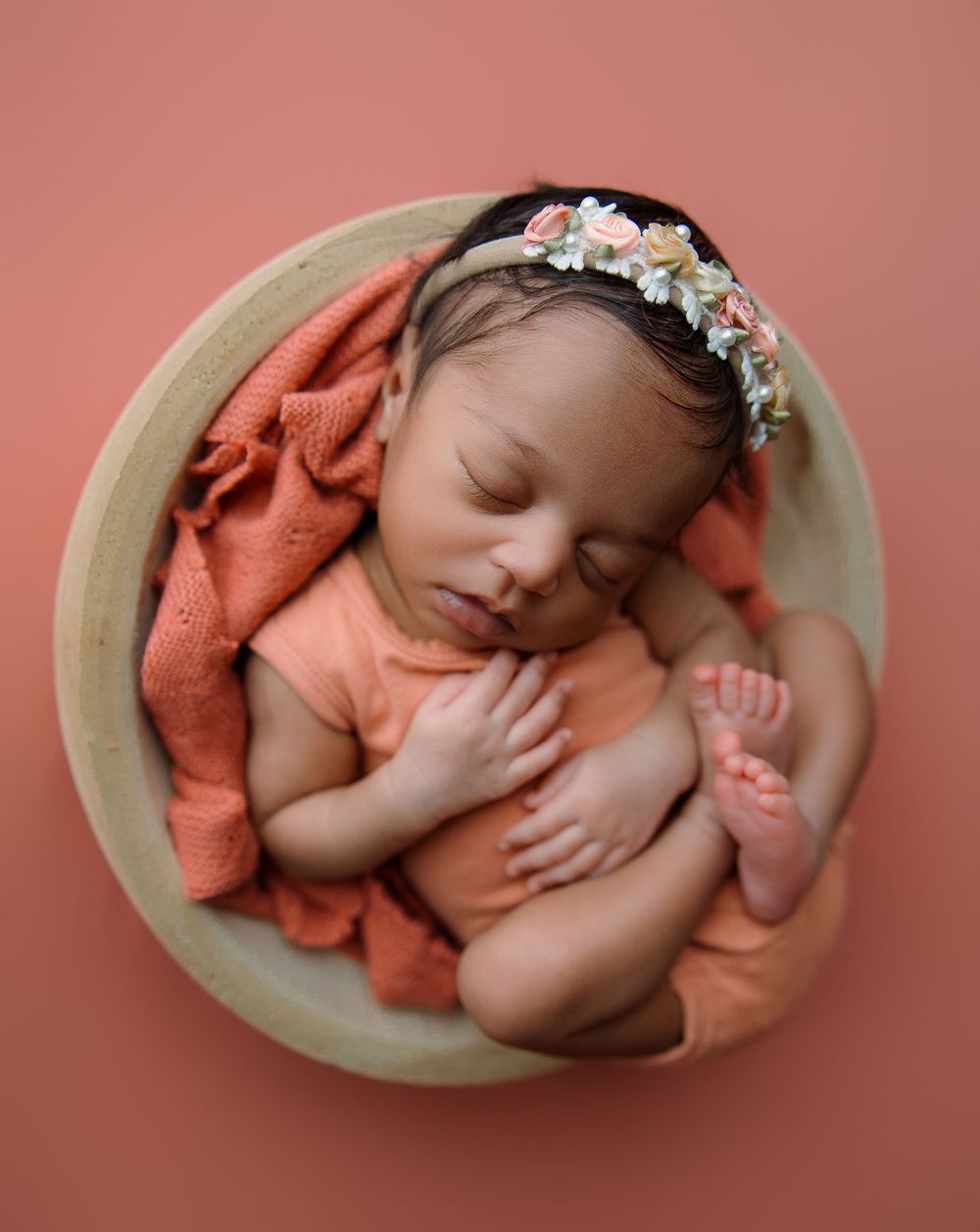 Sleeping newborn baby with floral headband resting in round bowl with coral-colored fabric on peachy background.