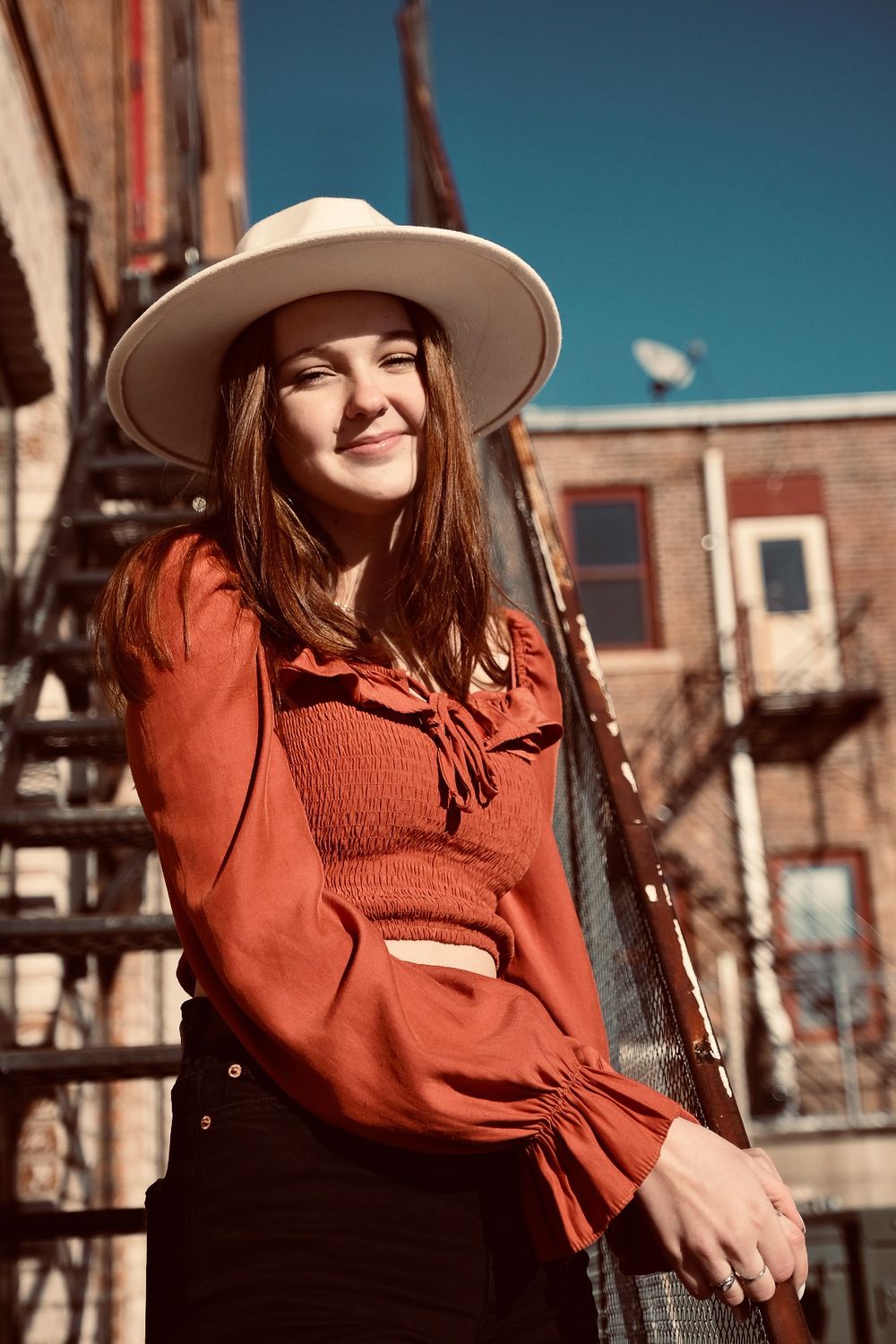 A person in a rust-colored sweater and wide-brimmed hat poses outdoors against an urban backdrop on a sunny day.