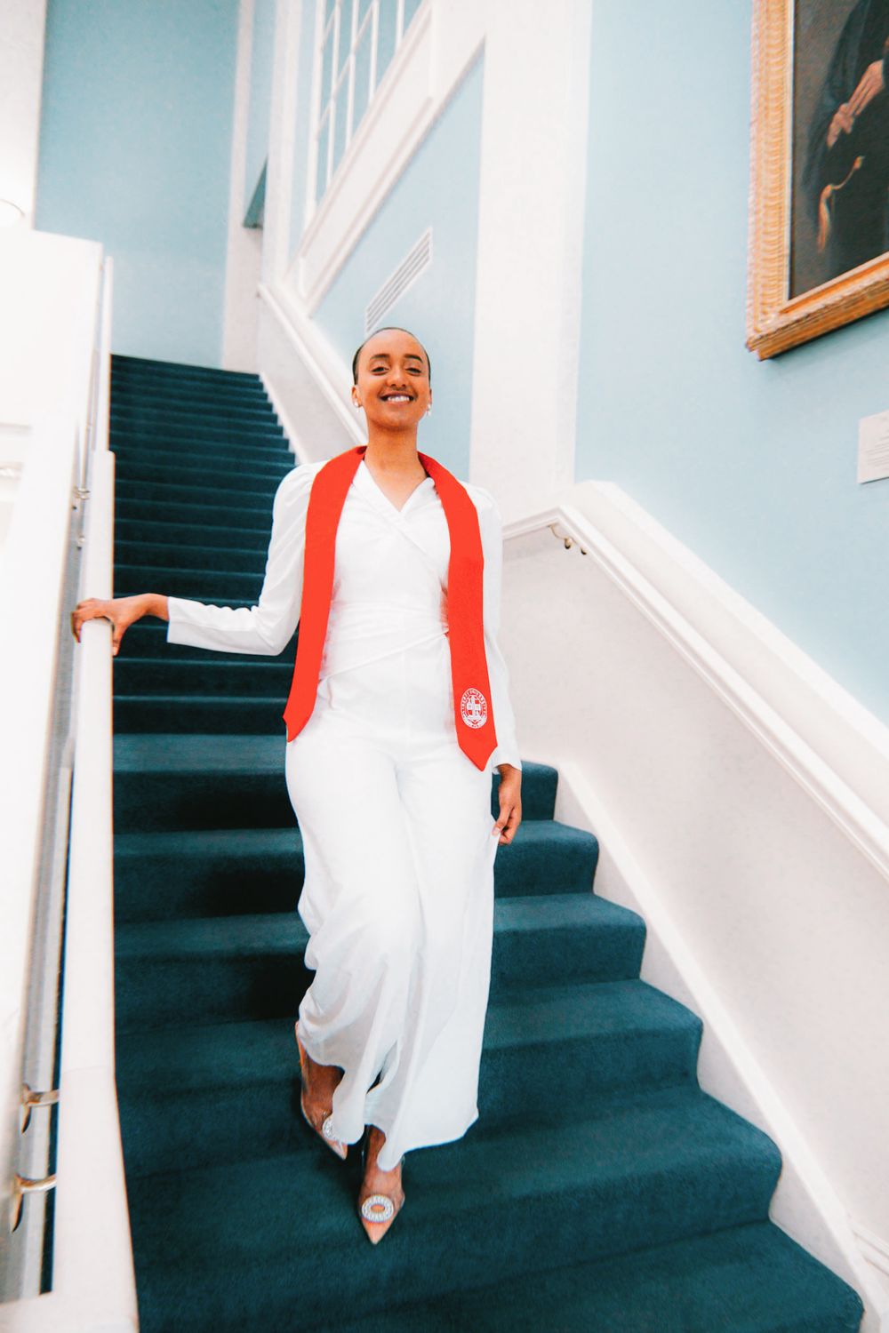 Someone wearing a red cardigan and white dress poses on teal carpeted stairs in an elegant interior setting.
